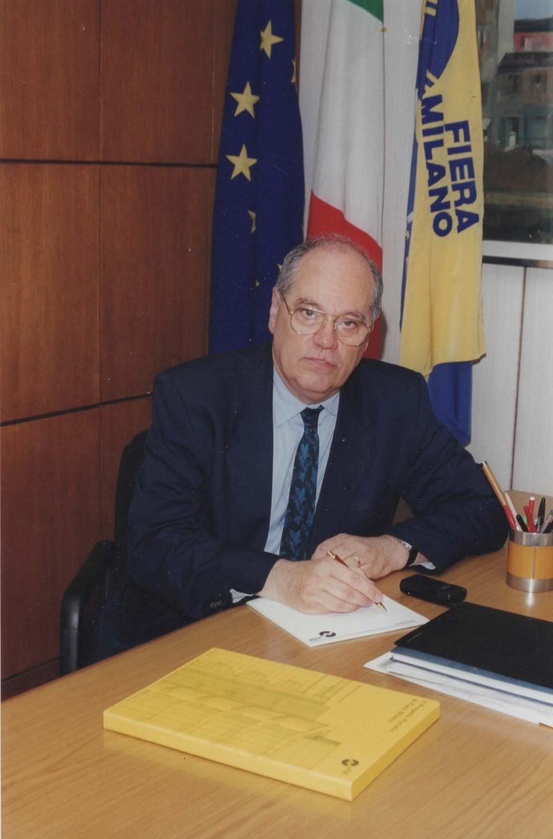 Peppino Zola alla scrivania, con le bandiere di Fiera Milano, dell'Italia e dell'Europa